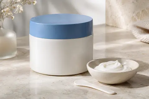 Pot blanc de baume hydratant à couvercle bleu posé sur une surface de pierre claire avec une spatule.