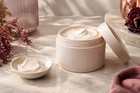 Crème hydratante pour le corps à la texture onctueuse, mise en scène sur pierre claire et céramique ivoire.