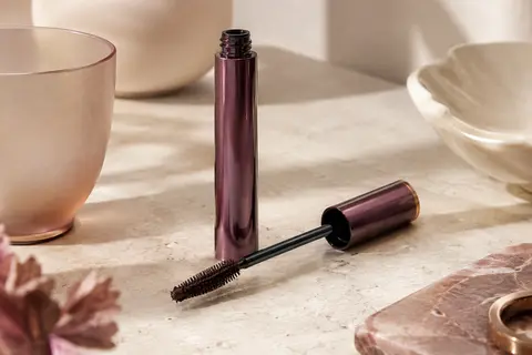 Mascara en tube avec sa brosse conique en fibres et une nuance brun noir sur une surface claire.