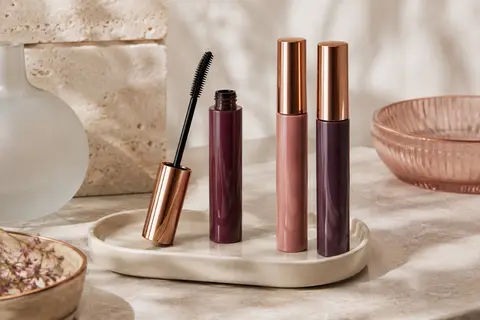 Trois tubes de mascara noir avec une brosse fuselée, posés sur une pierre claire près d’accessoires ivoire.