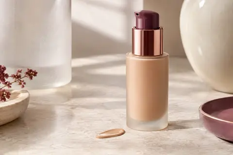 Flacon pompe de fond de teint fluide posé sur une pierre claire avec une touche de matière beige rosé