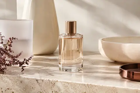 Flacon vaporisateur d’eau de parfum posé sur une pierre claire, près d’un verre dépoli et d’une céramique ivoire.