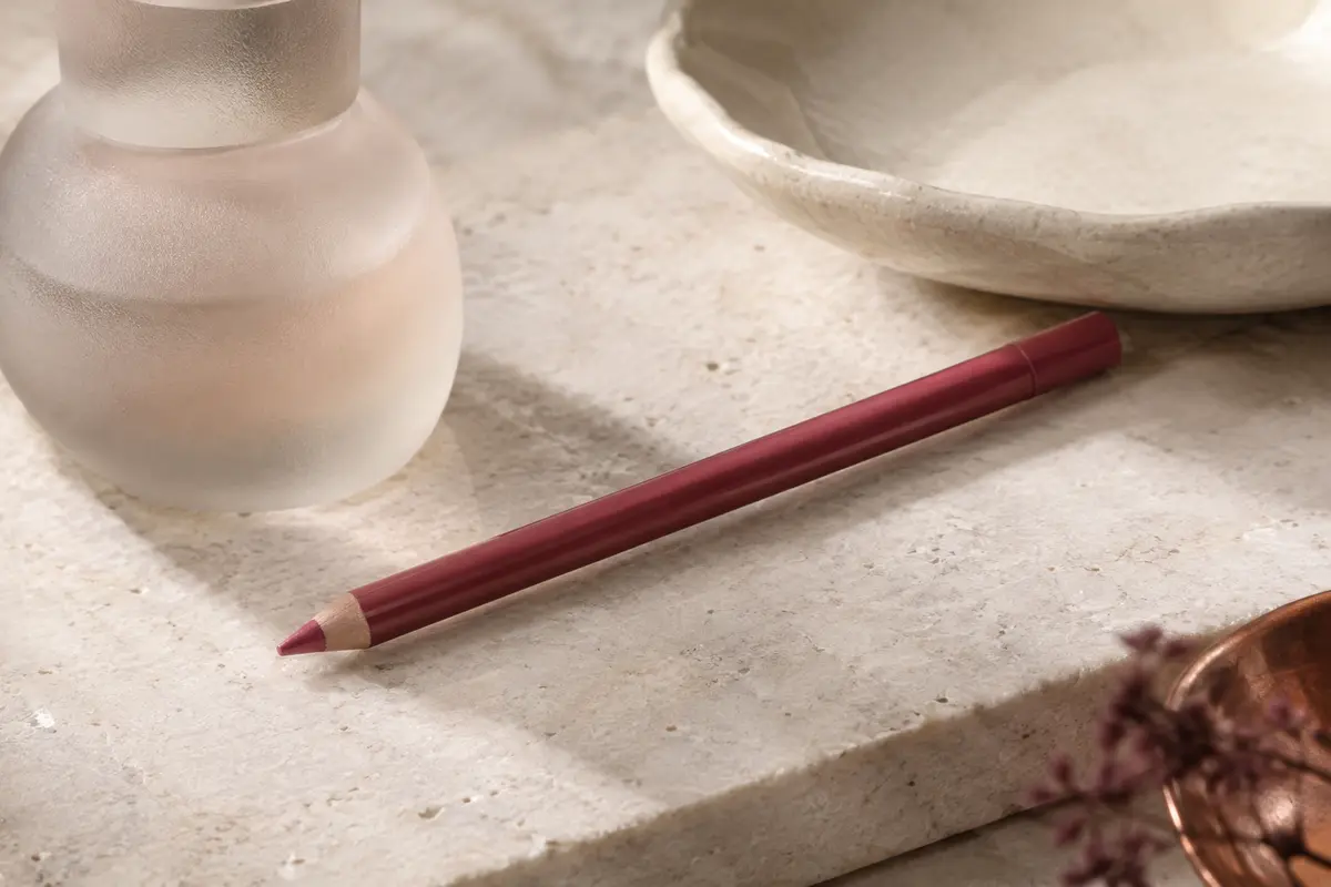 Crayon rouge à lèvres à mine fine posé sur une pierre claire, près d’un élément en céramique ivoire
