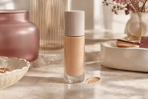 Flacon de fond de teint liquide beige sable posé sur une pierre claire, avec une touche de texture satinée.