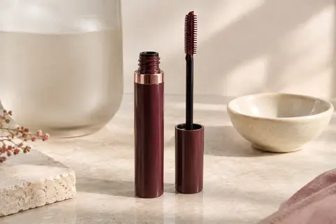 Tube de mascara liquide marron avec une brosse peigne posé sur une surface en pierre claire.