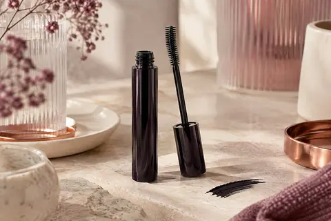 Mascara en tube avec brosse flexible et matière noire, posé sur une pierre claire près d’un verre dépoli.