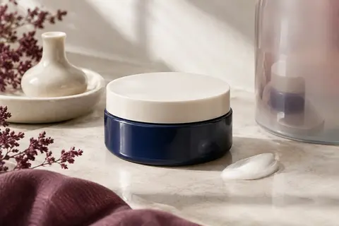 Pot ouvert de crème multi-usage posé sur une pierre claire avec une céramique ivoire et du verre dépoli.