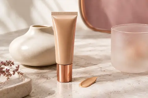 Tube de fond de teint liquide beige sable posé sur une pierre claire dans un décor épuré