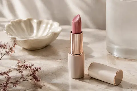Rouge à lèvres crème rose froid présenté ouvert sur une surface en pierre claire, près d’un élément en verre dépoli.